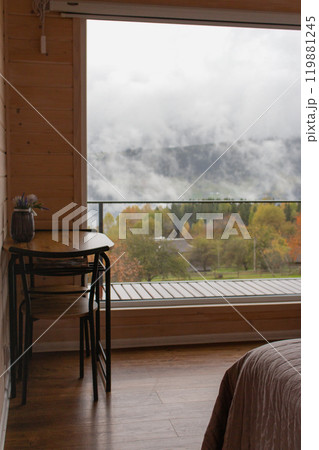 Table and chair on amazing foggy mountains background in the window. Silent autumn nature in mountains hotel. Room interior in tourist resort in Carpathians, Ukraine. Table and chair on amazing foggy mountains background in the window. Silent autumn nature in mountains hotel. Room interior in tourist resort in Carpathians, Ukraine. 119881245