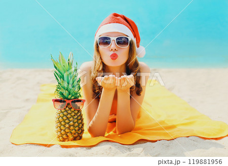 Christmas portrait woman in red santa hat with pineapple lying on beach blowing kiss on sea Christmas portrait woman in red santa hat with pineapple lying on beach blowing kiss on sea 119881956