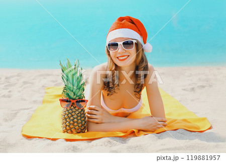 Christmas portrait young smiling woman in red santa hat and pineapple lying on beach on sea Christmas portrait young smiling woman in red santa hat and pineapple lying on beach on sea 119881957