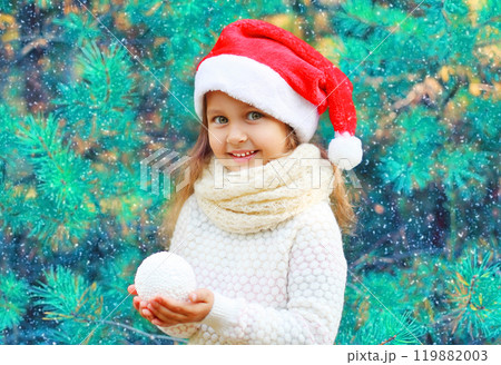 Christmas portrait smiling child little girl in santa red hat holding snowball near a branch tree 119882003