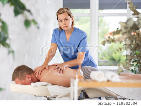 Female masseur performs back stroking and patting during massage session for male client Female masseur performs back stroking and patting during massage session for male client 119882304