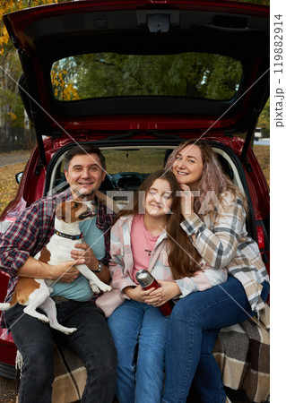 Happy parents with daughter and cute dog Jack Russel terrier sitting in car trunk on autumn day 119882914