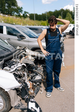 Full length vertical portrait of puzzled mechanic wondering looking at car bumper thinking of how to repair after accident during repairing car in repair workshop. Concept of professional car repair. Full length vertical portrait of puzzled mechanic wondering looking at car bumper thinking of how to repair after accident during repairing car in repair workshop. Concept of professional car repair. 119883641