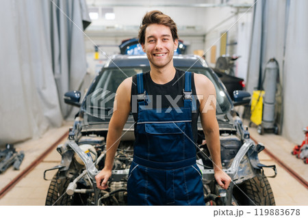Portrait of positive repairman in uniform standing smiling looking at camera in auto repair shop indoors. Confident professional talented mechanic male posing to camera, ready to repair vehicle. Portrait of positive repairman in uniform standing smiling looking at camera in auto repair shop indoors. Confident professional talented mechanic male posing to camera, ready to repair vehicle. 119883678