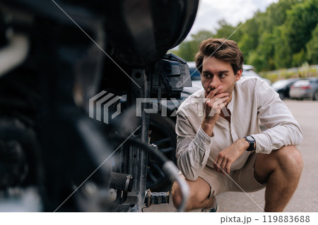 Frustrated young driver man assesses damage to car following accident on street, concerned about expenses for repairs and insurance. Worried car owner crouching next to damaged car in parking lot. 119883688