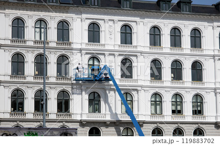Worker paints the facade of the building at heigt in lifting cradle. Construction workers at height working on crane in lifting bucket. 119883702