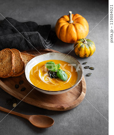 Pumpkin soup with cream, basil and seeds on a wooden board with fried bread on a dark background 119884021