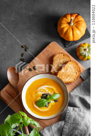 Pumpkin soup with cream, basil and seeds on a wooden board with fried bread, fresh herb and spoon  119884022
