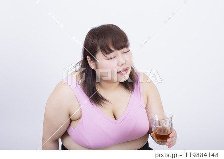 白い背景の前で美味しそうにコップの飲み物を飲むスポーツブラの太った若い女性 白い背景の前で美味しそうにコップの飲み物を飲むスポーツブラの太った若い女性 119884148