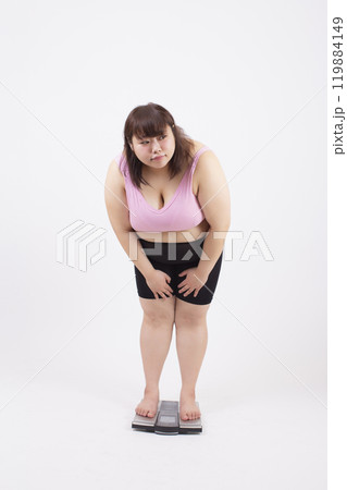 白い背景の前で周りを気にしながら体重計に乗るスポーツブラの太った若い女性 白い背景の前で周りを気にしながら体重計に乗るスポーツブラの太った若い女性 119884149