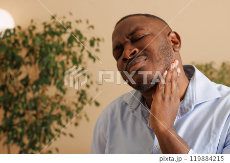 Man sitting on sofa in living room and feeling pain in neck and throat. Cozy home environment. Concept of discomfort, pain relief, home interior. Man sitting on sofa in living room and feeling pain in neck and throat. Cozy home environment. Concept of discomfort, pain relief, home interior. 119884215