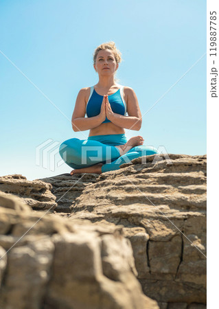 Yoga woman sitting in lotus position namaste hands cliff rock beach sea blue sky 119887785