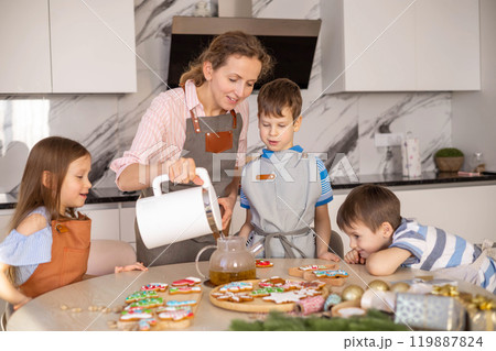 Happy young mother pouring tea into mugs. Tea party with Christmas gingerbread Happy young mother pouring tea into mugs. Tea party with Christmas gingerbread 119887824