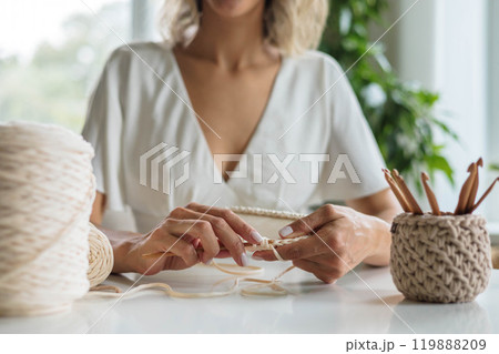 A young girl in a white dress sits at a table and knits a basket from beige yarn. 119888209