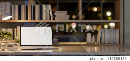 A laptop on a table by a bookshelf in a modern office at night, illuminated by a table lamp. 119889183