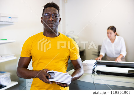 Happy middle-aged African American male typographer showing new notepads in the middle of the copy center Happy middle-aged African American male typographer showing new notepads in the middle of the copy center 119891905