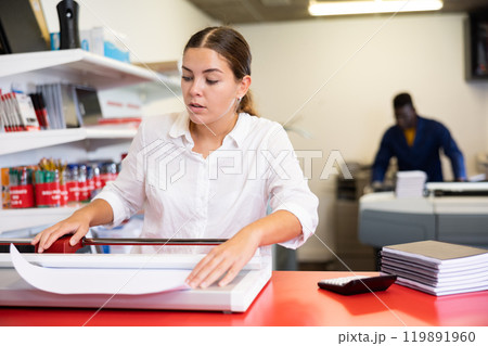 Concentrated young woman in white shirt using paper cutter on the table with planners and calculator in the printer house 119891960
