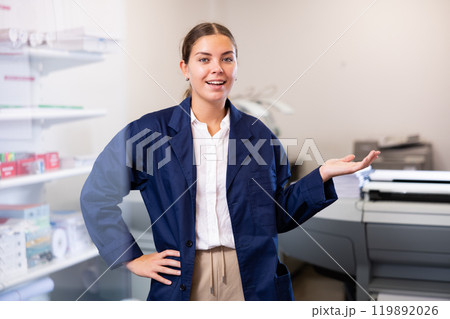 Cheerful young woman in blue robe uniform sticking out her left hand to show the workflow in the printer house Cheerful young woman in blue robe uniform sticking out her left hand to show the workflow in the printer house 119892026