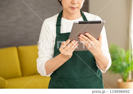 家のリビングでタブレットを見る緑のエプロンをしたシニア女性の主婦・家政婦 119896692