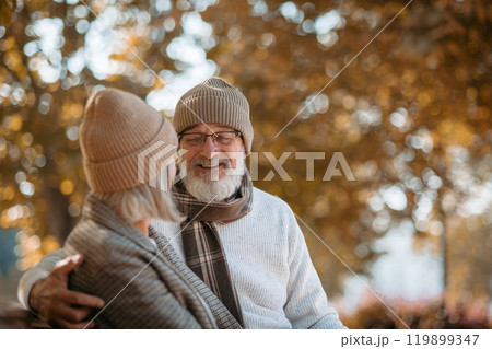 Portrait of beautiful senior couple sitting on bench in autumn park. Elderly husband and wife looking at each other lovingly. 119899347