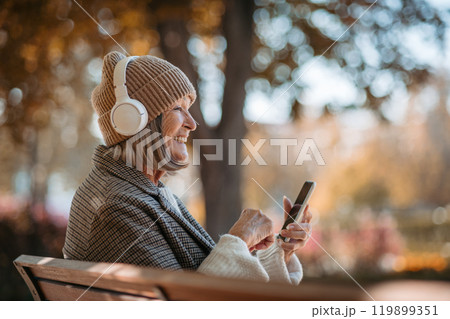 Portrait of a beautiful senior woman with headphones in autumn park, listening music and holding her phone. Portrait of a beautiful senior woman with headphones in autumn park, listening music and holding her phone. 119899351