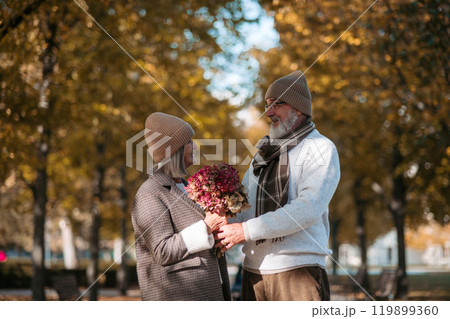 Elderly husband giving wife bouquet. Portrait of beautiful senior couple during walk in autumn park. 119899360