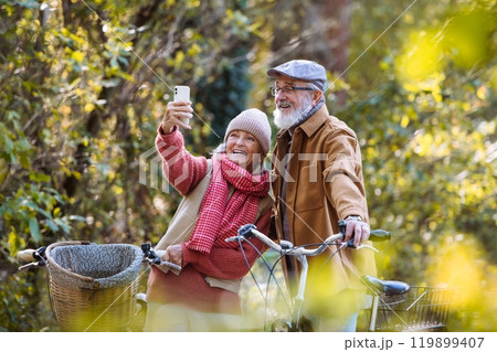 Lovely elderly couple on standing in the middle of autumn nature with their bikes, taking selfie with smartphone. 119899407