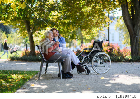 Elderly woman enjoying cup of coffee with her nurse in the park during warm autumn day. Young caregiver spending time with senior woman. Elderly woman enjoying cup of coffee with her nurse in the park during warm autumn day. Young caregiver spending time with senior woman. 119899419