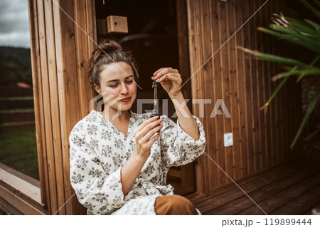 Beautiful woman doing her skin care routine outdoors, wearing bathrobe. Woman using facial skin serum with dropper. 119899444