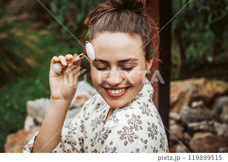 Beautiful woman massaging her face with rose quartz roller, sitting in garden. Outdoor skin care routine for young woman. Beautiful woman massaging her face with rose quartz roller, sitting in garden. Outdoor skin care routine for young woman. 119899515
