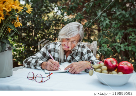 Beautiful older women is sitting in the garden and writing in her diary. Female senior is keeping daily journal, writing down thoughts and feelings. 119899592