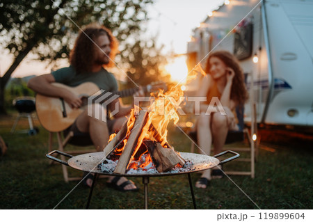 Couple sitting by campfire in the evening, enjoying peaceful moment. Man playing guitar and singing song to his girlfriend. Camping trip for young people. Couple sitting by campfire in the evening, enjoying peaceful moment. Man playing guitar and singing song to his girlfriend. Camping trip for young people. 119899604