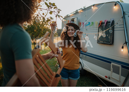 Young beautiful woman is on a camping trip in nature, putting lights on tree, preparing for evening. 119899616