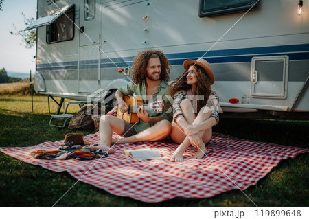 Couple sitting by caravan, enjoying peaceful moment. Man playing guitar and singing song to his girlfriend. Camping trip for young people. 119899648