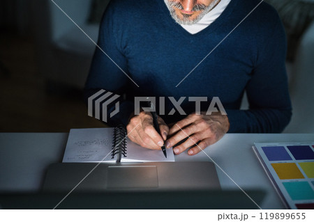 Close up of man working from home office at evening, writing notes in planner. Telecommuting and home office. Close up of man working from home office at evening, writing notes in planner. Telecommuting and home office. 119899655