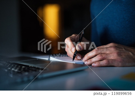 Close up of man working from home office at evening, writing notes in planner. Telecommuting and home office. Close up of man working from home office at evening, writing notes in planner. Telecommuting and home office. 119899656