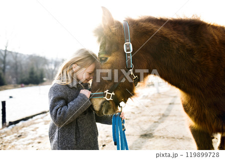 Young girl spending cold winter weekend on family farm, taking care of her brown fluffy pony, hugging him. Young girl spending cold winter weekend on family farm, taking care of her brown fluffy pony, hugging him. 119899728