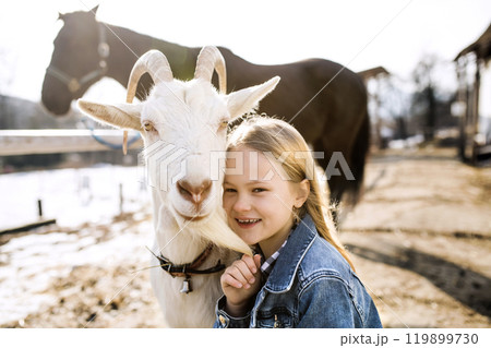 Young girl spending cold winter weekend on a farm, taking care of animals and petting white goat. Young girl spending cold winter weekend on a farm, taking care of animals and petting white goat. 119899730