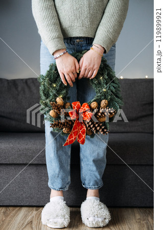 Person holding handmade Christmas wreath with pinecones and red bow, standing in cozy home setting. DIY holiday decor, festive minimalism, and winter crafts. 119899921