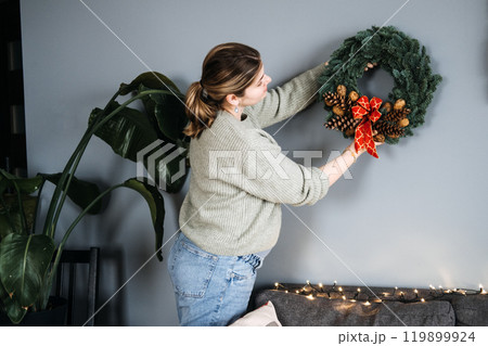 Woman hanging handmade Christmas wreath with pinecones and red ribbon. DIY holiday decor adds a personal touch to festive home decorating. Woman hanging handmade Christmas wreath with pinecones and red ribbon. DIY holiday decor adds a personal touch to festive home decorating. 119899924