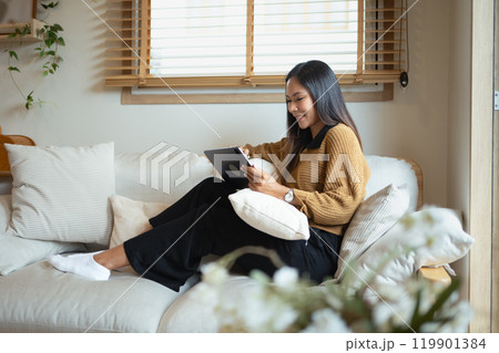 Charming young adult woman using digital tablet while relaxing on a cozy sofa Charming young adult woman using digital tablet while relaxing on a cozy sofa 119901384