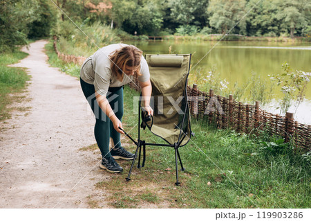 30s young woman preparing her chair amidst the beauty of the nature. Camping, tourism and travel concept. A happy blonde woman explores a forest and lake. Full body 119903286