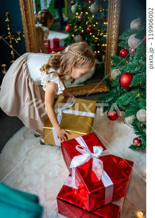 Little girl taking a present out from under the Christmas tree with red, gold and silver toys and garlands. Christmas and New Year at home Little girl taking a present out from under the Christmas tree with red, gold and silver toys and garlands. Christmas and New Year at home 119906002