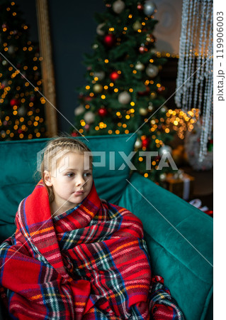 Little girl sitting wrapped in a warm red blanket against the background of a Christmas tree with red golden toys and a glowing garland at home 119906003