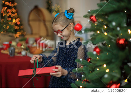 A joyful little girl in stylish glasses and a beautiful festive dress stands beside a sparkling Christmas tree, cradling a beautifully wrapped gift in her hands. moment to unwrap her present 119906204