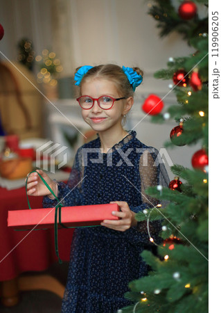 A joyful little girl in stylish glasses and a beautiful festive dress stands beside a sparkling Christmas tree, cradling a beautifully wrapped gift in her hands. moment to unwrap her present 119906205