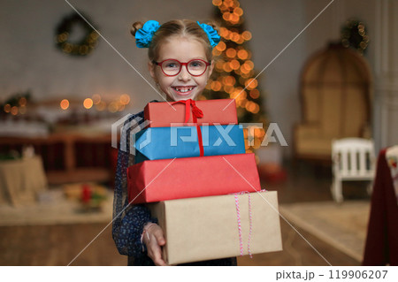 A joyful little girl in stylish glasses and a beautiful festive dress stands beside a sparkling Christmas tree, cradling a beautifully wrapped gift in her hands. moment to unwrap her present A joyful little girl in stylish glasses and a beautiful festive dress stands beside a sparkling Christmas tree, cradling a beautifully wrapped gift in her hands. moment to unwrap her present 119906207