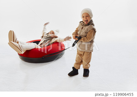 Happy, smiling young boy, dressed in beige winter clothing, pulling red snow tube with his sister on it. Excitement of wintertime activities. Happy, smiling young boy, dressed in beige winter clothing, pulling red snow tube with his sister on it. Excitement of wintertime activities. 119906455