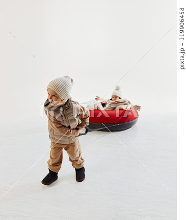 Little boy in winter clothing pulling his sister on red snow tube, showcasing joy and warmth of family activities during winter. Little boy in winter clothing pulling his sister on red snow tube, showcasing joy and warmth of family activities during winter. 119906458