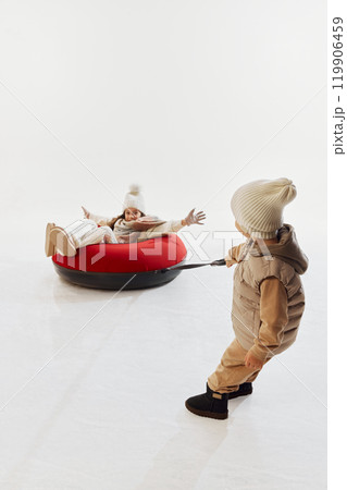 Young boy in warm winter clothes, pulling his sister on red snow tube across smooth snowy surface. Young boy in warm winter clothes, pulling his sister on red snow tube across smooth snowy surface. 119906459